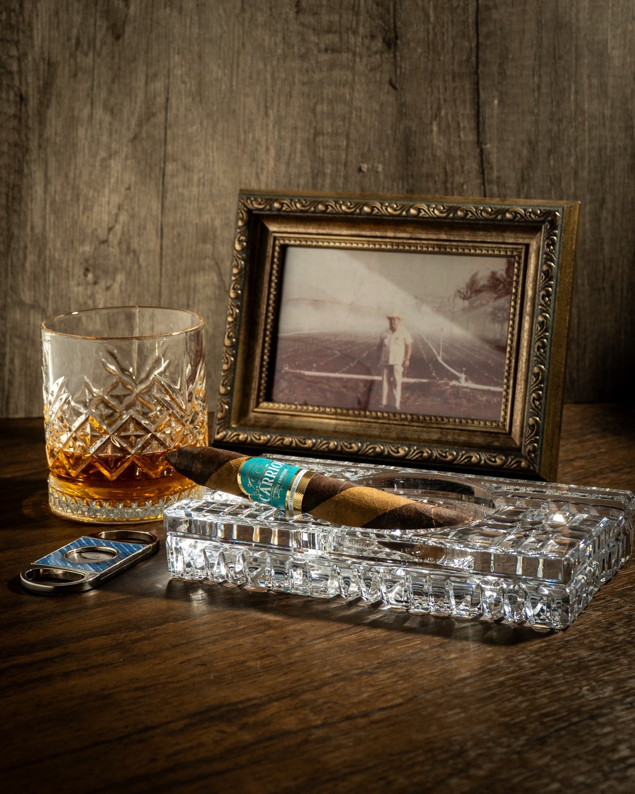 Fotografía antigua de un hombre en un campo de tabaco, junto a un puro Carrion y un vaso de whisky
