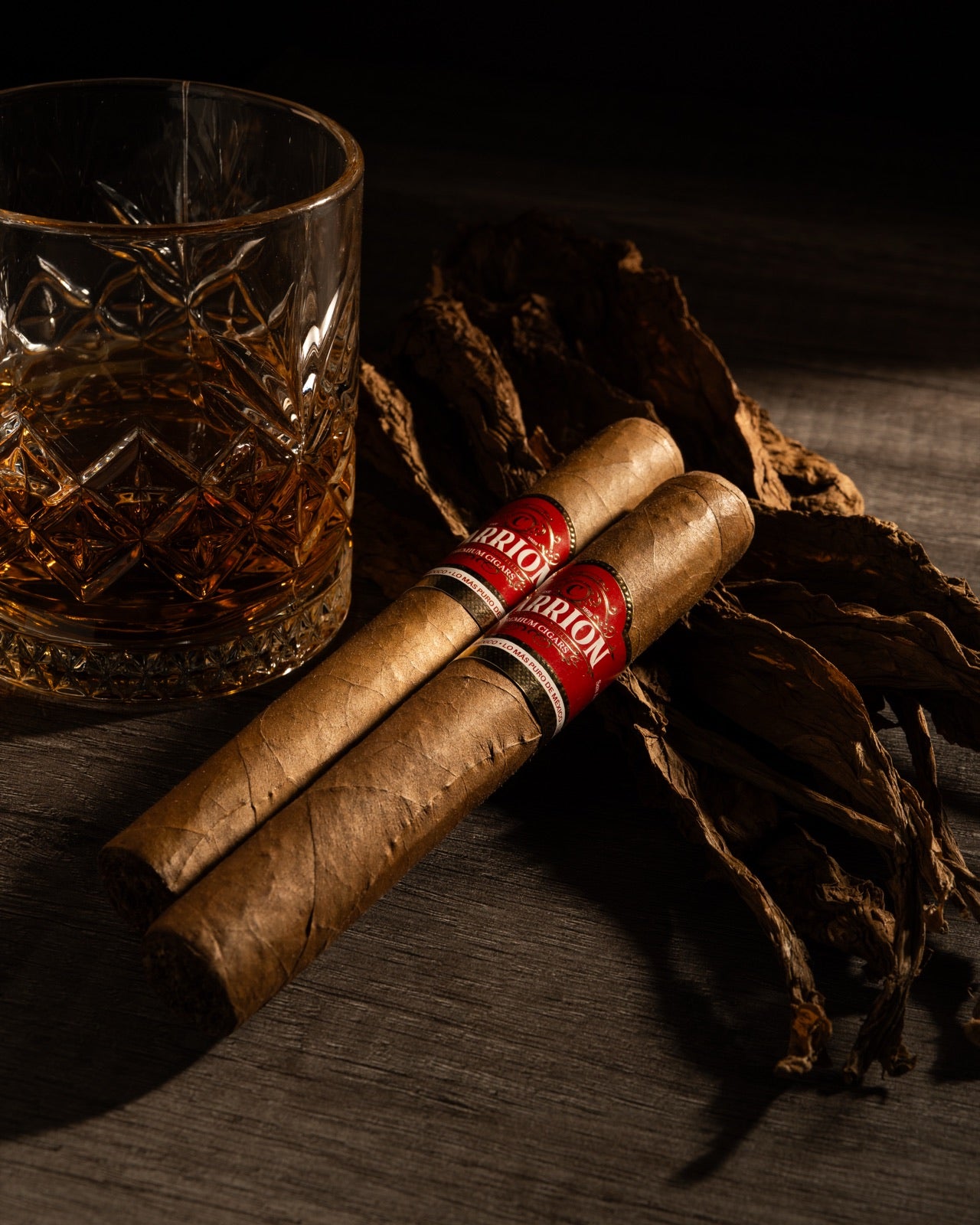 Dos puros Carrion sobre hojas de tabaco curado, junto a un vaso de whisky